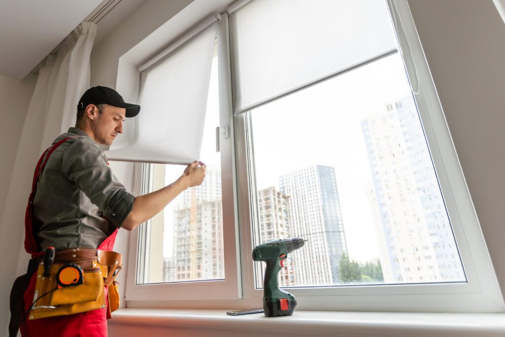 Worker in uniform installing roller window blind indoors, focus on drill Professional Window Covering Installation